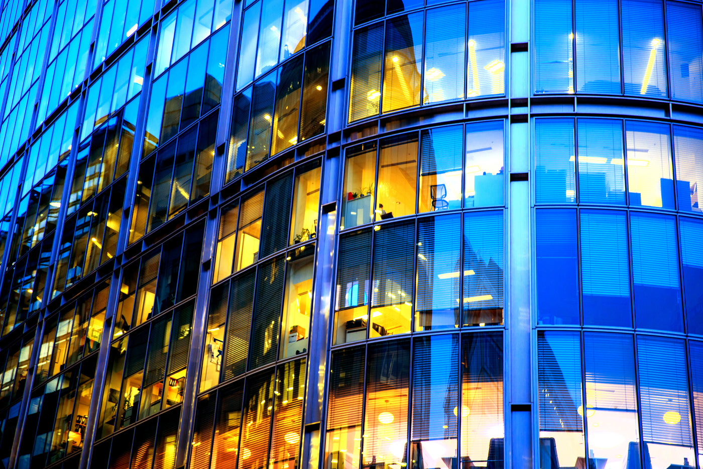 Framsida av modern kontorsbyggnad, gul färg från ljuskällor, armaturer och led-lysrör i olika nyanser som syns genom fönster. Även blå nyans återspeglas på fönsterpartierna. Blått och gult är även färgerna i Lampsson logotype.