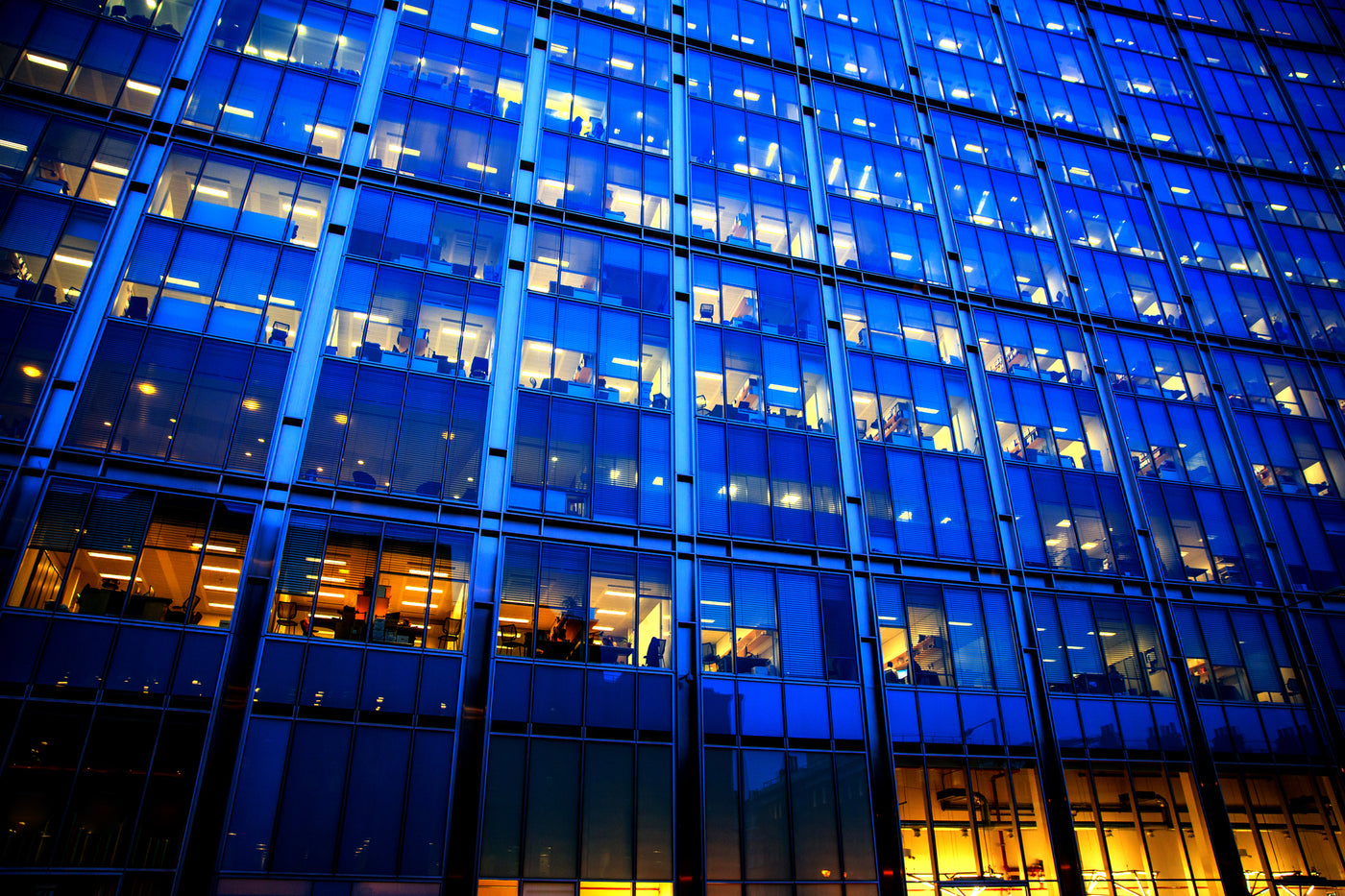 Framsida av modern kontorsbyggnad med stora fönster och med synliga järnbalkar mellan de stora fönsterpartierna. Det lyser gult från armaturer, lysrör och lampor i olika nyanser från flertalet rum samtidigt som glaspartierna återspeglar mörkblå himmel. Det gula från ljuskällor och blåa från himlen är även färgerna i lampssons logotype.