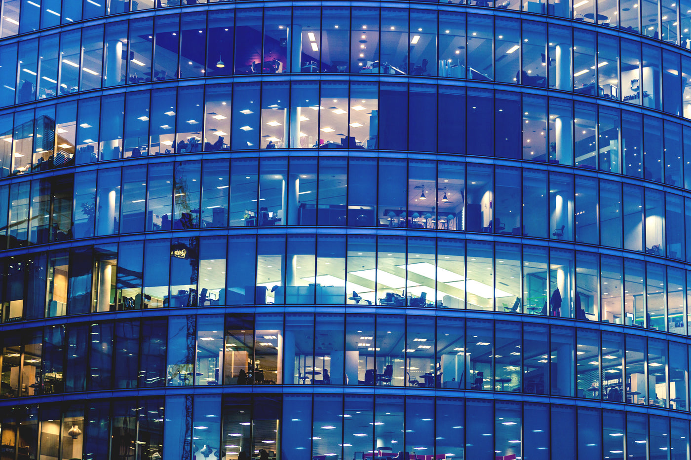 Framsida modern kontorsbyggnad. Genom de stora fönsterpartierna lyser det gult i olika nyanser från armaturer, ledbelysning och lampor. Även blåa partier syns på fönsterpartierna. Färgerna blått och gult är även färgerna i Lampsson logotype.