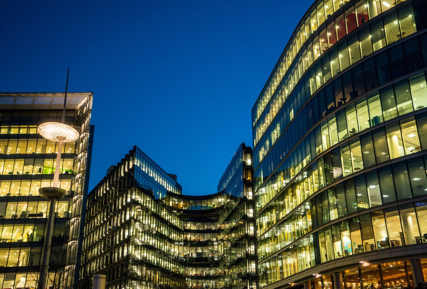 Kontorsbyggnader med mörkblå himmel i bakgrunden. Det lyser gult i olika nyanser genom fönsterpartierna från ledarmaturer, led ljuskällor och led belysning. Blått och gult är även färgerna i Lampssons logotype.